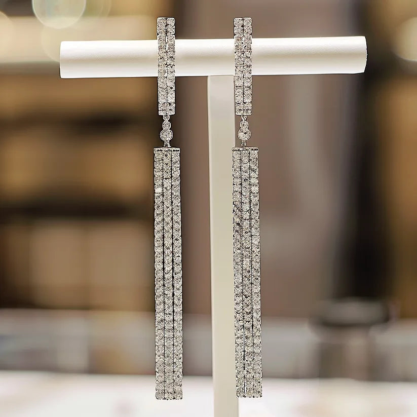 The image shows a pair of elegant, long dangling earrings on a white stand. Each earring has a diamond-encrusted vertical bar at the top, connected to multiple strands of diamond-encrusted bars hanging vertically. 