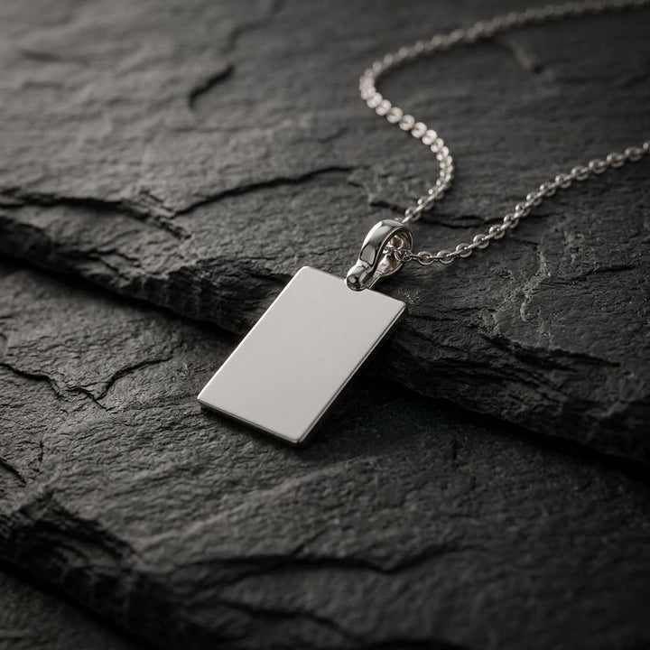 White gold plain rectangular tag pendant, 19 x 13 mm, 0.5 mm thick, on a white gold chain resting on dark slate stone