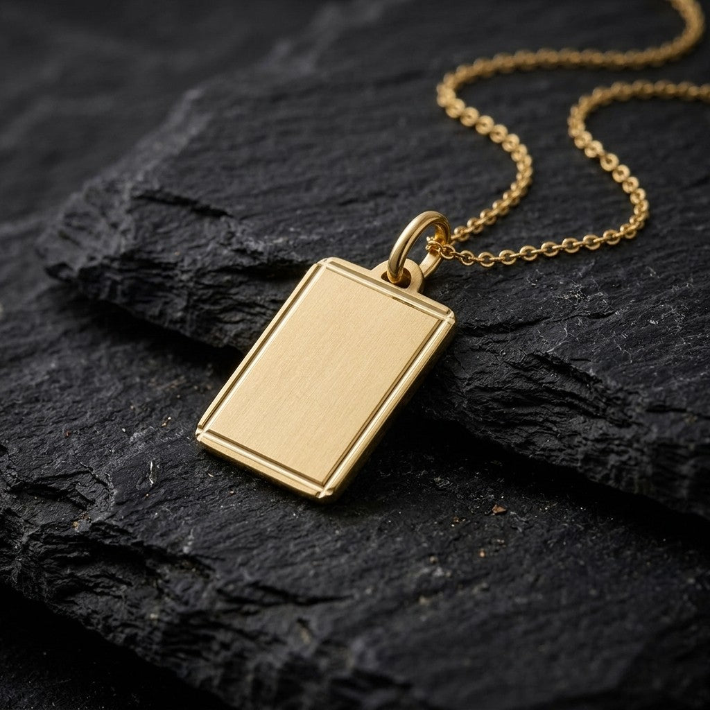 18K yellow gold satin finish rectangular tag pendant with polished line border, 19.4 x 14.9 mm, on a yellow gold chain resting on dark slate stone