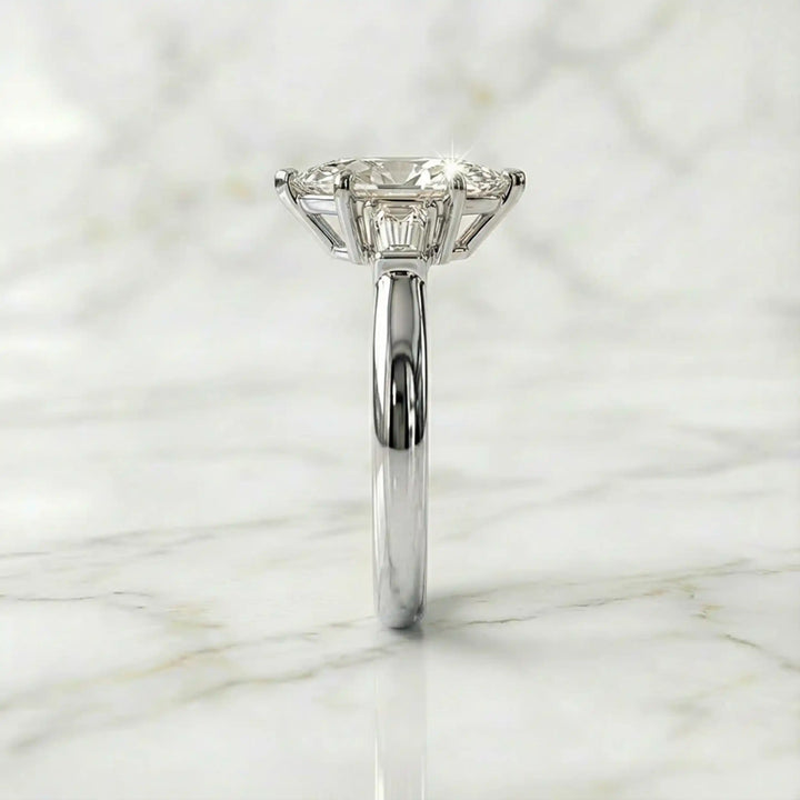 Three-stone marquise lab-grown diamond engagement ring displayed on marble surface.
