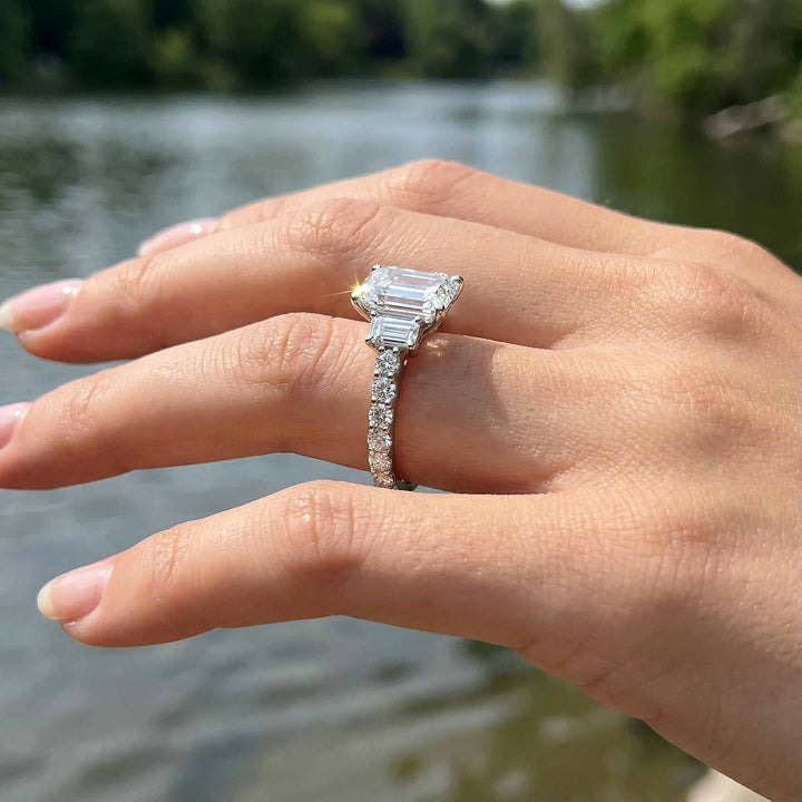 Emerald cut three stone diamond engagement ring with pavé band on display.
