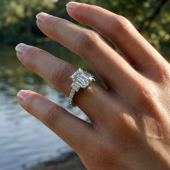 Emerald cut three stone diamond engagement ring with pavé band on display.

