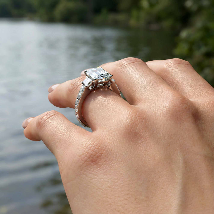 Emerald cut three stone diamond engagement ring with pavé band on display.
