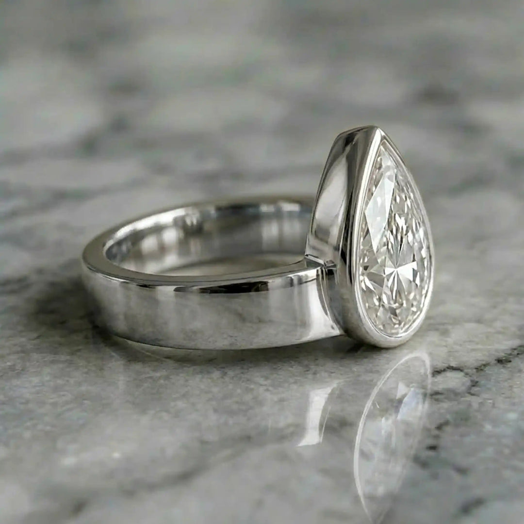 3 carat pear-shaped lab-grown diamond bezel solitaire engagement ring displayed on marble surface.
