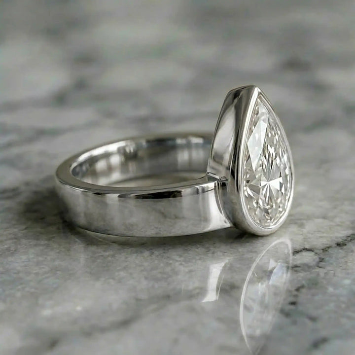 3 carat pear-shaped lab-grown diamond bezel solitaire engagement ring displayed on marble surface.
