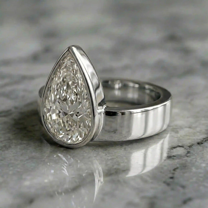 3 carat pear-shaped lab-grown diamond bezel solitaire engagement ring displayed on marble surface.
