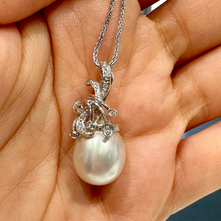 A close-up image of a pendant featuring a large South Sea pearl, suspended from an ornate silver setting adorned with small gemstones. The setting is designed with elegant, swirling motifs that add a decorative flourish around the pearl, which displays a lustrous sheen with subtle iridescent colors. The pendant hangs from a fine silver chain.