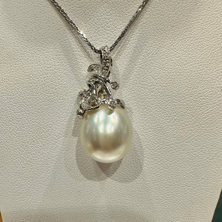 A close-up image of a pendant featuring a large South Sea pearl, suspended from an ornate silver setting adorned with small gemstones. The setting is designed with elegant, swirling motifs that add a decorative flourish around the pearl, which displays a lustrous sheen with subtle iridescent colors. The pendant hangs from a fine silver chain.