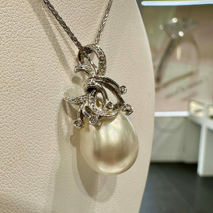 A close-up image of a pendant featuring a large South Sea pearl, suspended from an ornate silver setting adorned with small gemstones. The setting is designed with elegant, swirling motifs that add a decorative flourish around the pearl, which displays a lustrous sheen with subtle iridescent colors. The pendant hangs from a fine silver chain.