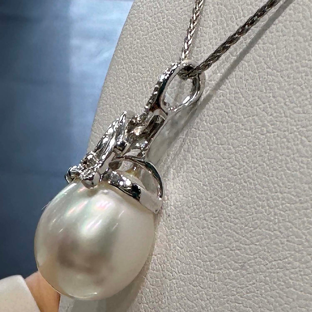 A close-up image of a pendant featuring a large South Sea pearl, suspended from an ornate silver setting adorned with small gemstones. The setting is designed with elegant, swirling motifs that add a decorative flourish around the pearl, which displays a lustrous sheen with subtle iridescent colors. The pendant hangs from a fine silver chain.