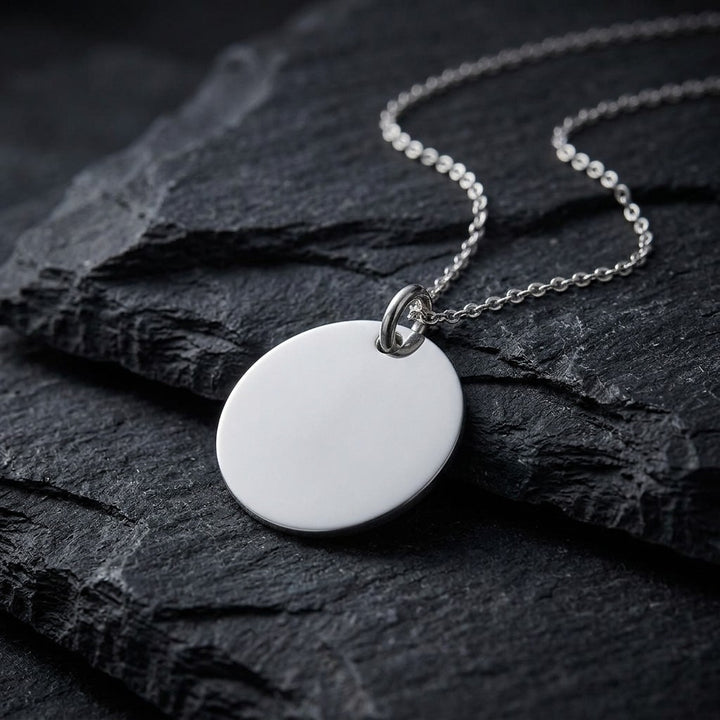 Sterling silver round disc tag pendant with a polished face and jump ring, 22.5 mm, on a sterling silver chain resting on dark slate stone