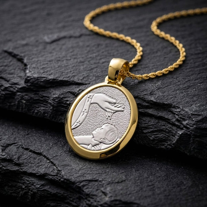 Two-tone gold rounded oval baptism medal pendant with a polished yellow gold frame around a white gold stippled panel showing a raised hand pouring water over a sleeping infant beside a priest's stole with crosses, 16 mm wide, on a gold chain resting on dark slate stone