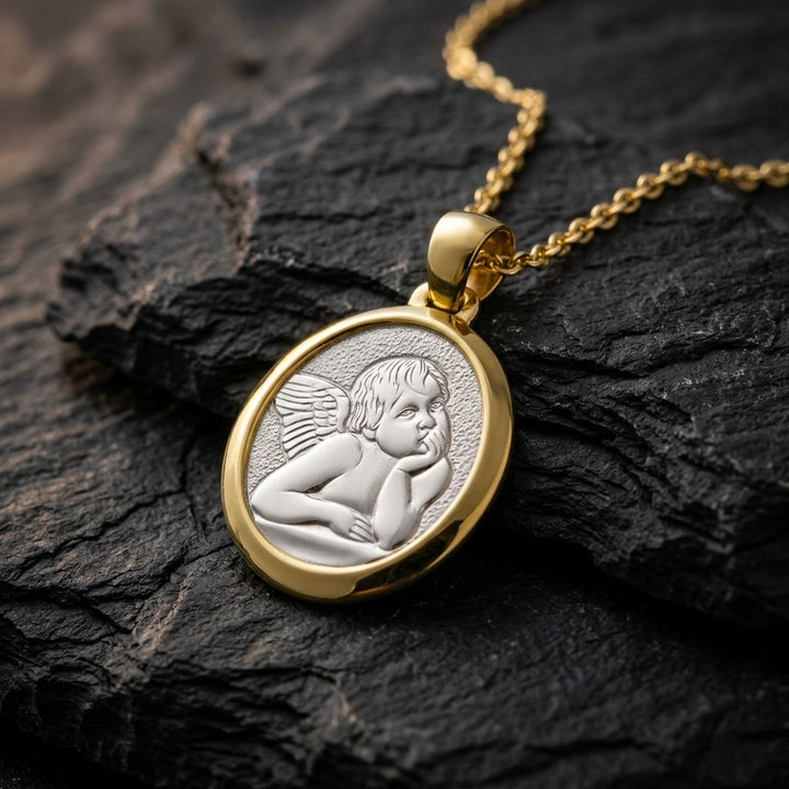 Two-tone gold rounded oval angel medal pendant with a raised white gold Raphael cherub in relief in a polished yellow gold frame, 19.9 x 16 mm, on a yellow gold chain resting on dark slate stone
