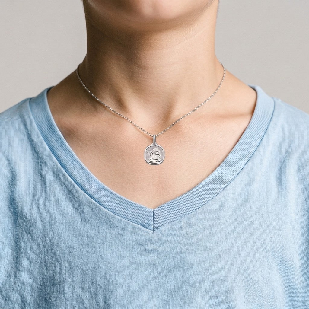 White gold cushion-shaped angel medal pendant with a raised Raphael cherub in relief on a stippled background, 14.6 mm, worn by a boy on a fine white gold chain