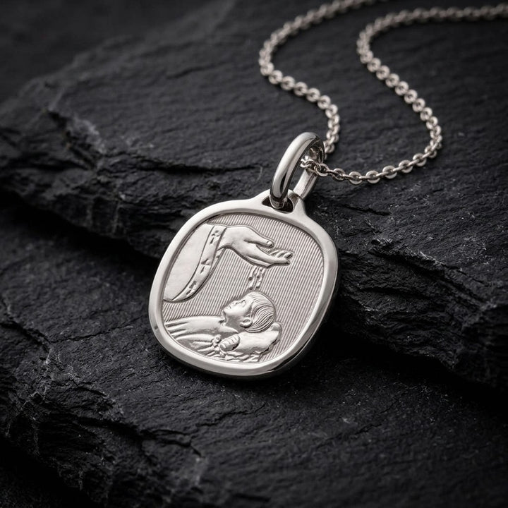 White gold cushion square baptism medal pendant with a polished rim, ribbed background and a raised hand pouring water over a sleeping infant beside a priest's stole with crosses, 15 mm wide, on a white gold chain resting on dark slate stone