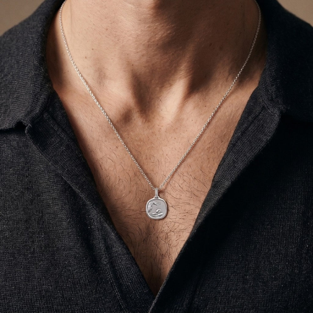 White gold cushion square baptism medal pendant with a polished rim, ribbed background and a raised hand pouring water over a sleeping infant beside a priest's stole with crosses, 15 mm wide, worn by a man on a fine white gold chain