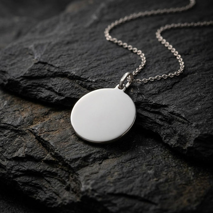 White gold polished round tag pendant, 18 mm diameter, 0.5 mm thick, on a white gold chain resting on dark slate stone