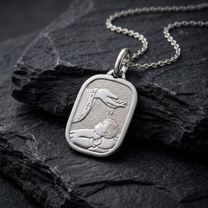 White gold rounded rectangle baptism medal pendant with a wide polished rim, stippled background and a raised hand pouring water over a sleeping infant beside a priest's stole with crosses, 14.4 mm wide, on a white gold chain resting on dark slate stone