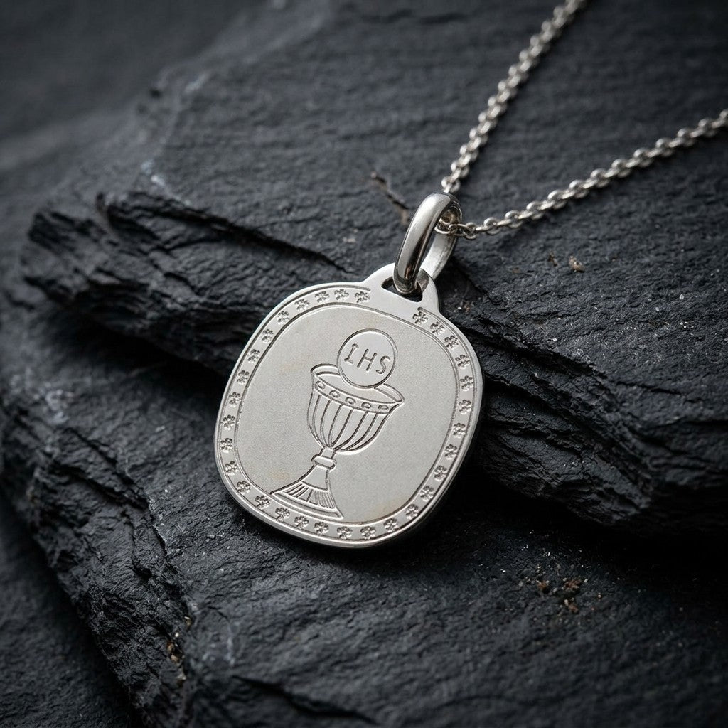 White gold rounded square communion medal pendant with an engraved chalice and host inside a clover-patterned border, 16.2 mm wide, on a white gold chain resting on dark slate stone