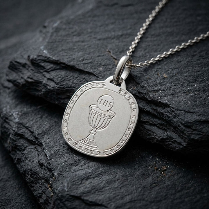 White gold rounded square communion medal pendant with an engraved chalice and host inside a clover-patterned border, 16.2 mm wide, on a white gold chain resting on dark slate stone