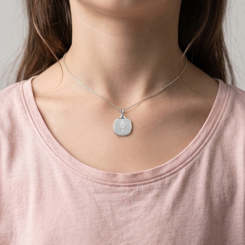 White gold rounded square communion medal pendant with an engraved chalice and host inside a clover-patterned border, 16.2 mm wide, worn by a girl on a fine white gold chain
