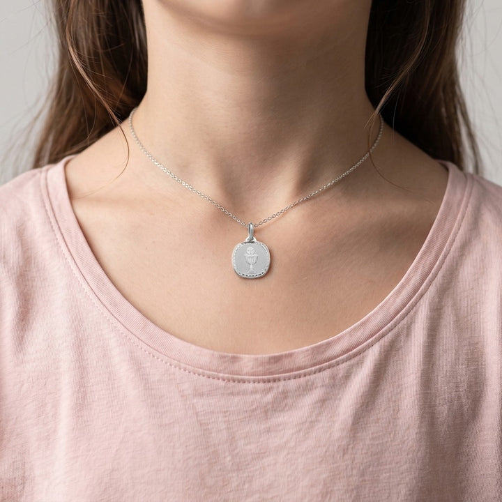 White gold rounded square communion medal pendant with an engraved chalice and host inside a clover-patterned border, 16.2 mm wide, worn by a girl on a fine white gold chain