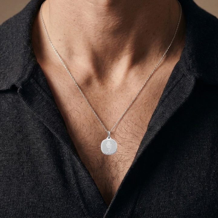 White gold rounded square communion medal pendant with an engraved chalice and host inside a clover-patterned border, 16.2 mm wide, worn by a man on a fine white gold chain