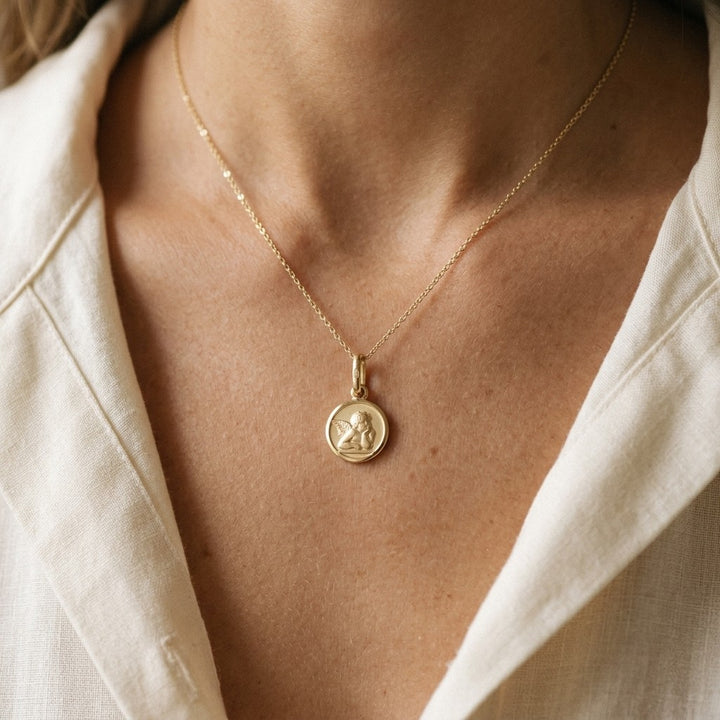 Yellow gold round angel medal pendant with a raised Raphael cherub in relief, 10 mm, worn by a woman on a fine yellow gold chain