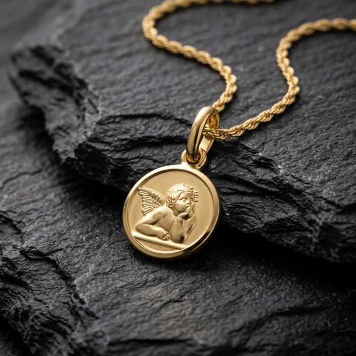 Yellow gold round angel medal pendant with a raised Raphael cherub in relief, 12 mm, on a yellow gold chain resting on dark slate stone