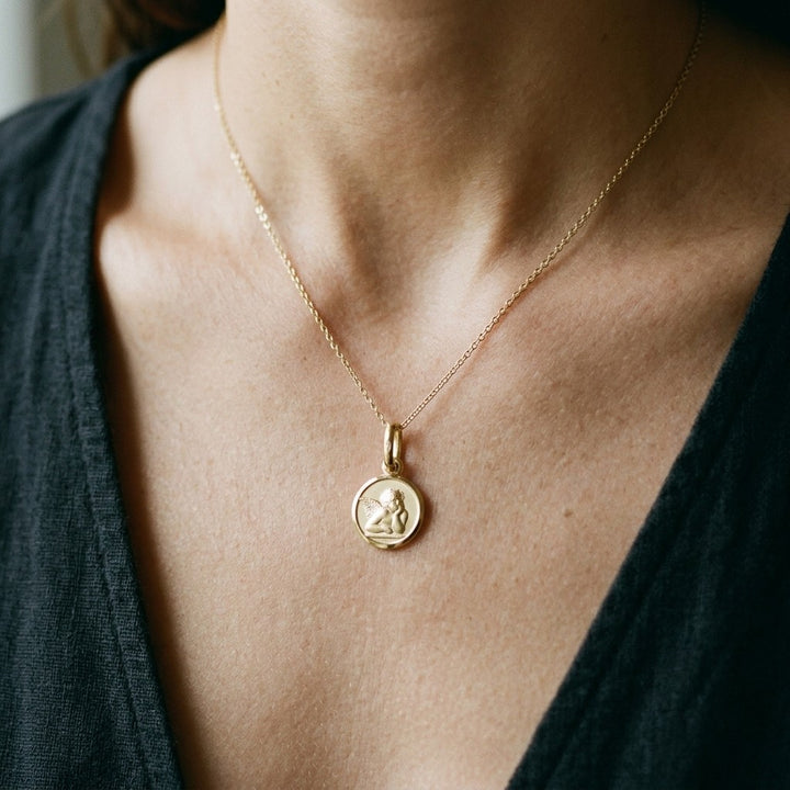 Yellow gold round angel medal pendant with a raised Raphael cherub in relief, 12 mm, worn by a woman on a fine yellow gold chain