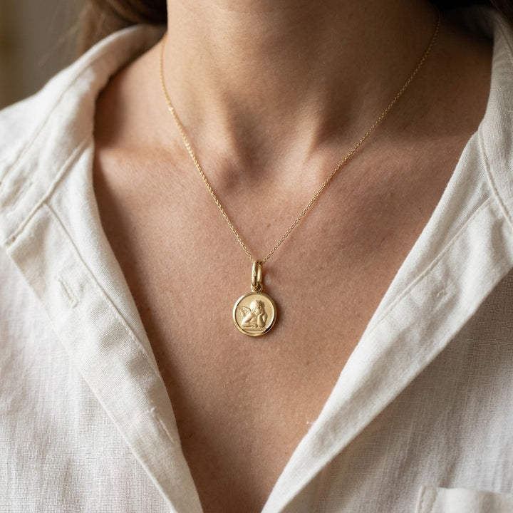 Yellow gold round angel medal pendant with a raised Raphael cherub in relief, 16 mm, worn by a girl on a fine yellow gold chain