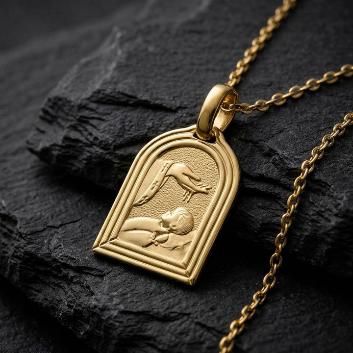 Yellow gold arched church-window baptism medal pendant with a double polished border and a raised hand pouring water over a sleeping infant beside a priest's stole with crosses, 14 mm wide, on a yellow gold chain resting on dark slate stone