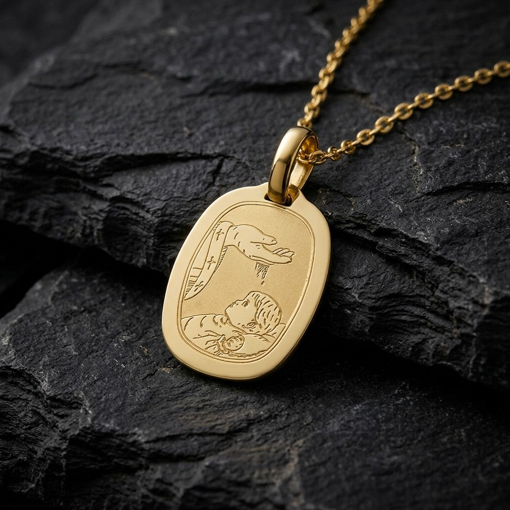 Yellow gold barrel-shaped rounded octagon baptism medal pendant with a wide polished rim and a fine engraved scene of a hand pouring water over a sleeping infant beside a priest's stole with crosses, 16 mm wide, on a yellow gold chain resting on dark slate stone