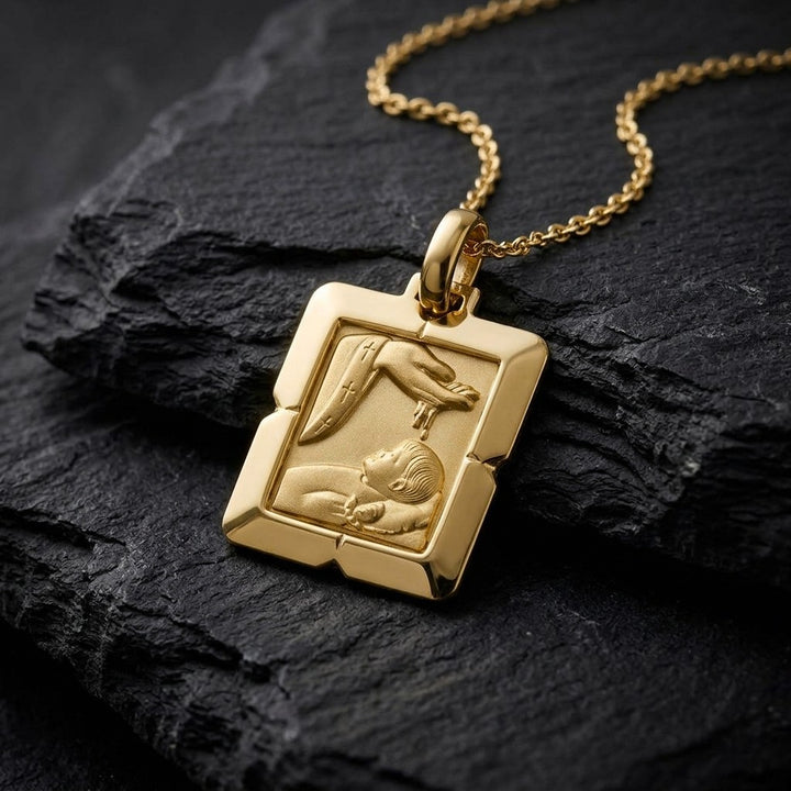 Yellow gold bevelled square baptism medal pendant with stepped corners and a raised hand pouring water over a sleeping infant beside a priest's stole with crosses, 17 mm wide, on a yellow gold chain resting on dark slate stone