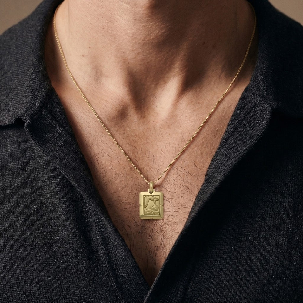 Yellow gold bevelled square baptism medal pendant with stepped corners and a raised hand pouring water over a sleeping infant beside a priest's stole with crosses, 17 mm wide, worn by a man on a fine yellow gold chain