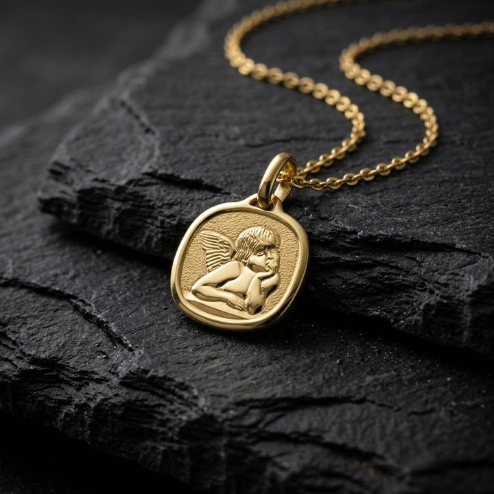 Yellow gold cushion-shaped angel medal pendant with a raised Raphael cherub in relief on a stippled background, 14.6 mm, on a yellow gold chain resting on dark slate stone