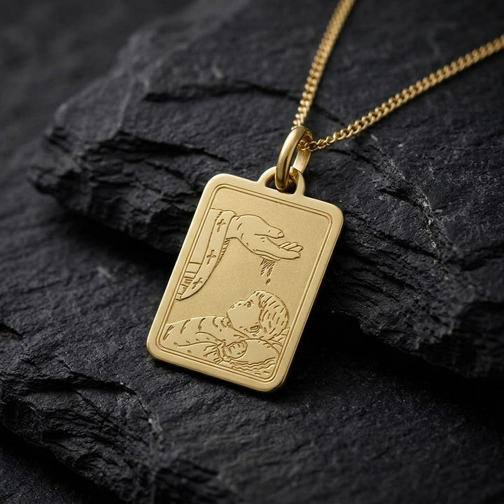Yellow gold rectangular baptism medal pendant with rounded corners and a fine engraved scene of a hand pouring water over a sleeping infant beside a priest's stole with crosses, 15 mm wide, on a yellow gold chain resting on dark slate stone