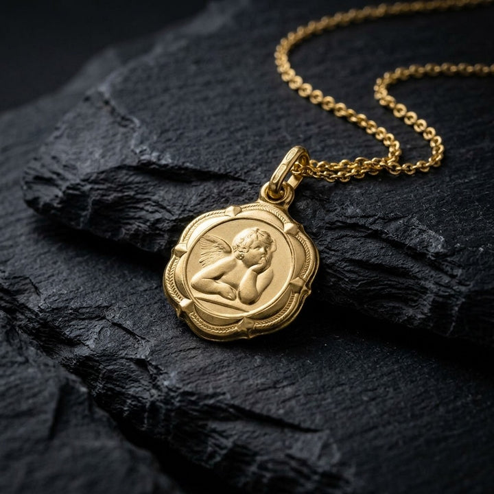Yellow gold four-lobed angel medal pendant with a raised Raphael cherub in relief and beaded corner ornaments, 14 mm, on a yellow gold chain resting on dark slate stone