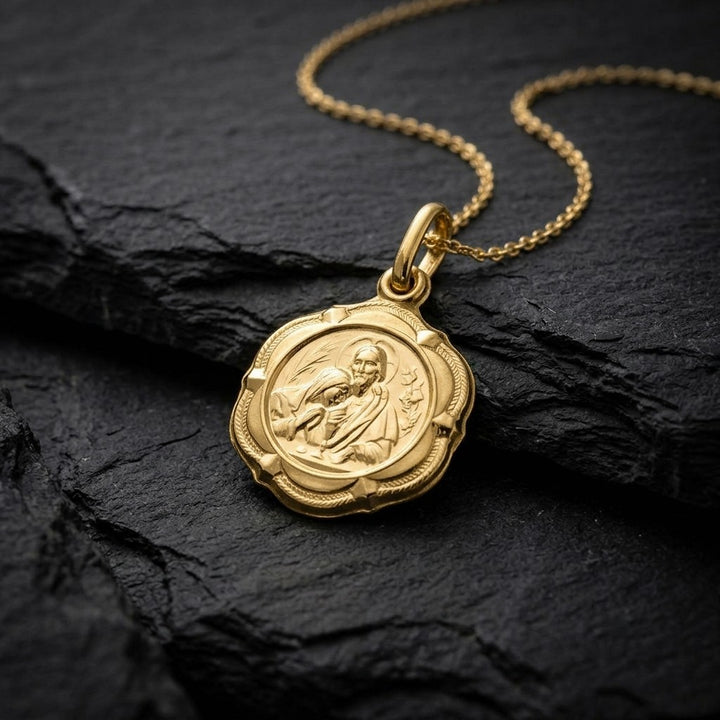 Yellow gold four-lobed communion medal pendant with a braided rim and four corner studs around a round centre of Jesus with a halo offering the communion host to a kneeling child, with wheat ears on the left and grapes on the right, 14 mm wide, on a yellow gold chain resting on dark slate stone
