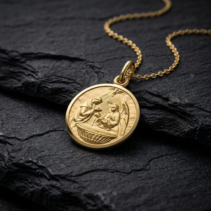 Yellow gold round baptism medal pendant with a raised priest, angel and infant at the font scene and a smooth polished rim, 16 mm, on a yellow gold chain resting on dark slate stone
