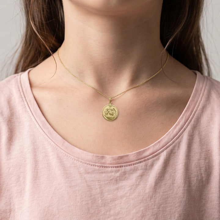 Yellow gold round baptism medal pendant with a raised priest, angel and infant at the font scene and a smooth polished rim, 16 mm, worn by a girl on a fine yellow gold chain