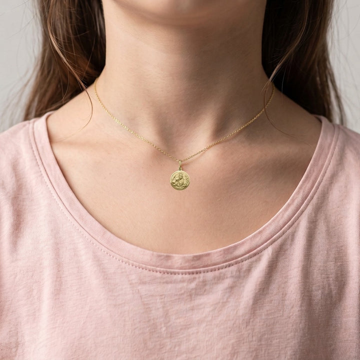 Yellow gold round communion medal pendant with a raised polished rim around a satin field of Jesus with a halo offering the communion host to a kneeling child, with wheat ears on the left and grapes on the right, 12 mm wide, worn by a girl on a fine yellow gold chain