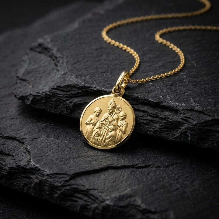 Yellow gold round confirmation medal pendant with a bishop in a tall mitre laying his hand on two kneeling children, standing children at each side and a polished rim, 12 mm wide, on a yellow gold chain resting on dark slate stone