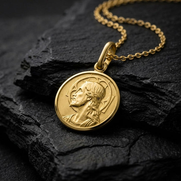 Yellow gold round Jesus medal pendant with a raised profile bust of Jesus facing left with long wavy hair and a beard, a halo behind the head and a polished rim, 12 mm wide, on a yellow gold chain resting on dark slate stone