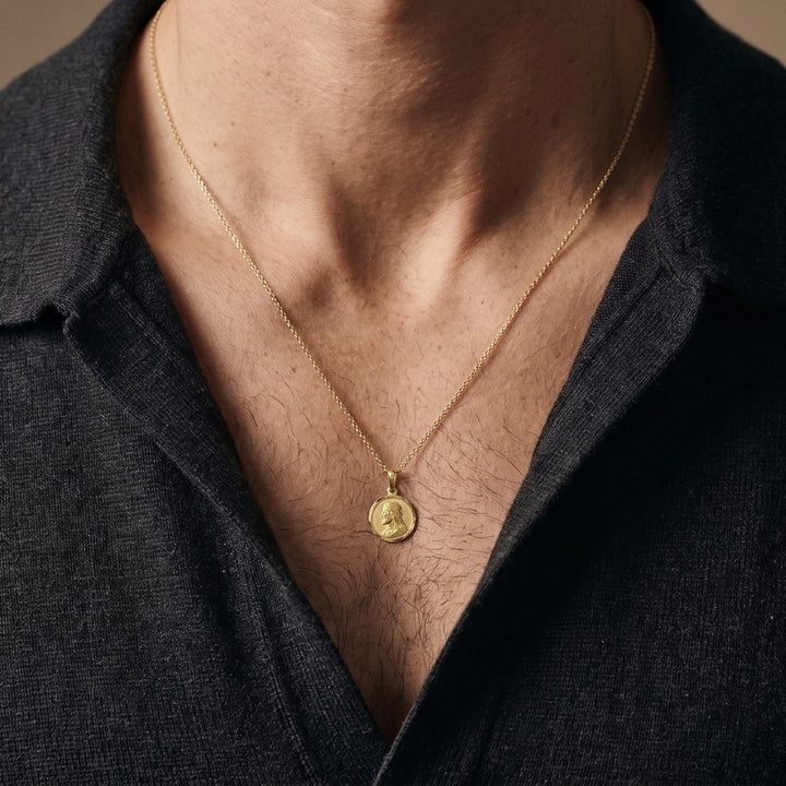Yellow gold round Jesus medal pendant with a raised profile bust of Jesus facing left with long wavy hair and a beard, a halo behind the head and a polished rim, 12 mm wide, worn by a man on a fine yellow gold chain