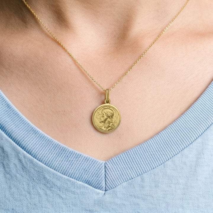 Yellow gold round Jesus medal pendant with a raised profile bust of Jesus facing left with long wavy hair and a beard, a halo behind the head and a polished rim, 14 mm wide, worn by a boy on a fine yellow gold chain, close-up view