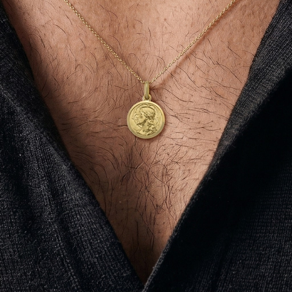 Yellow gold round Jesus medal pendant with a raised profile bust of Jesus facing left with long wavy hair and a beard, a halo behind the head and a polished rim, 14 mm wide, worn by a man on a fine yellow gold chain, close-up view