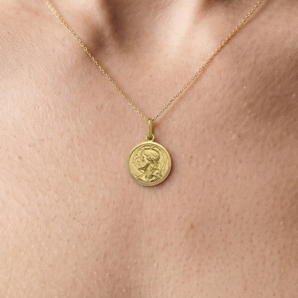 Yellow gold round Jesus medal pendant with a raised profile bust of Jesus facing left with long wavy hair and a beard, a halo behind the head and a polished rim, 14 mm wide, worn by a woman on a fine yellow gold chain, close-up view