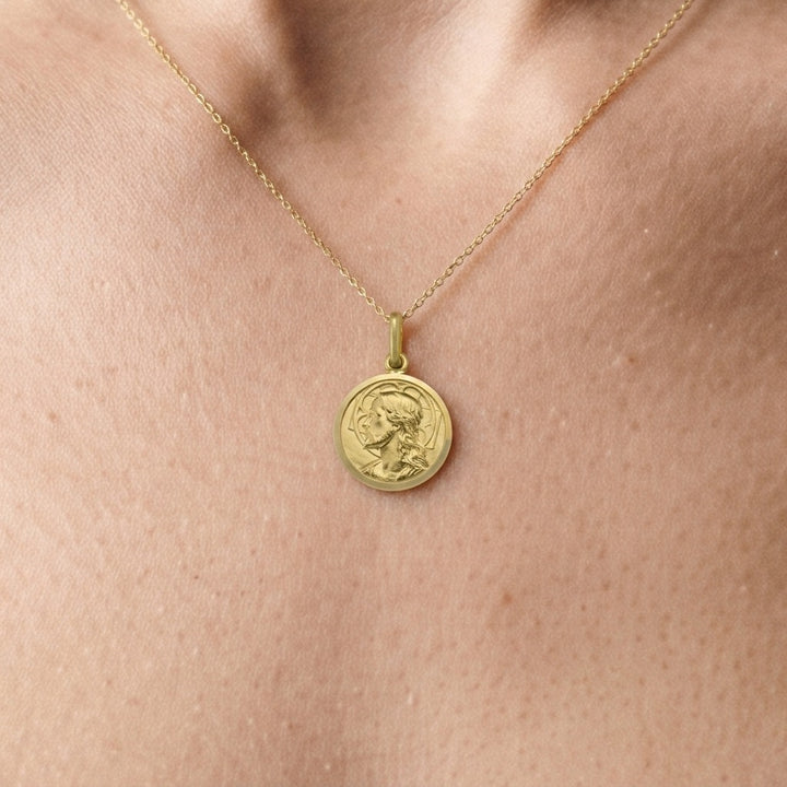 Yellow gold round Jesus medal pendant with a raised profile bust of Jesus facing left with long wavy hair and a beard, a halo behind the head and a polished rim, 14 mm wide, worn by a woman on a fine yellow gold chain, close-up view