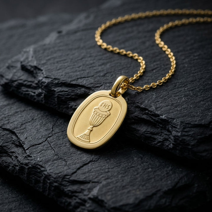 Yellow gold rounded octagon communion medal pendant with an engraved chalice and host, 16 mm wide, on a yellow gold chain resting on dark slate stone